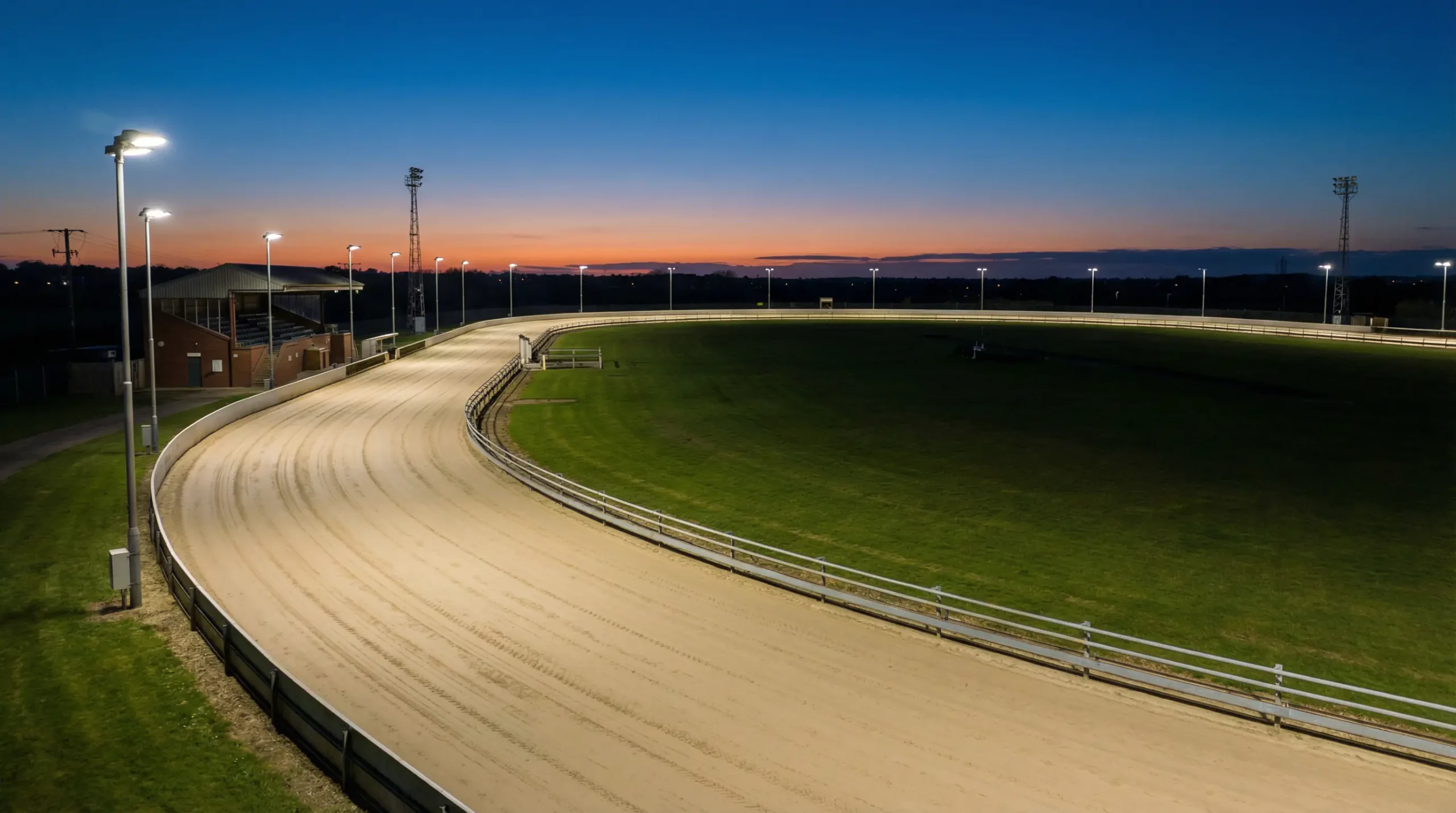 Aerial view of a full oval UK greyhound sand track at dusk with floodlights on